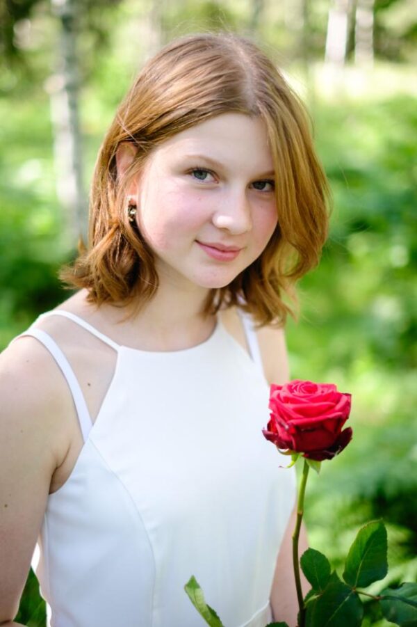 A young girl in the forest during her graduation photoshoot, holding a red rose and facing the camera.