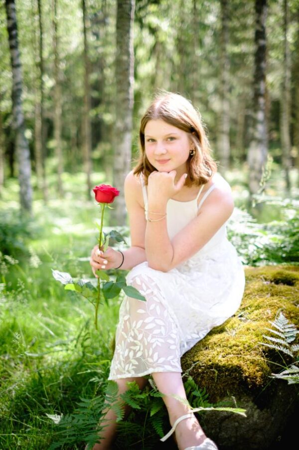 A young girl is in the forest during her graduation photo shoot, sitting on a mossy rock and looking directly at the camera.