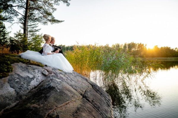 A wedding couple is sitting on a rock in front of the sea, gazing at the sunset.