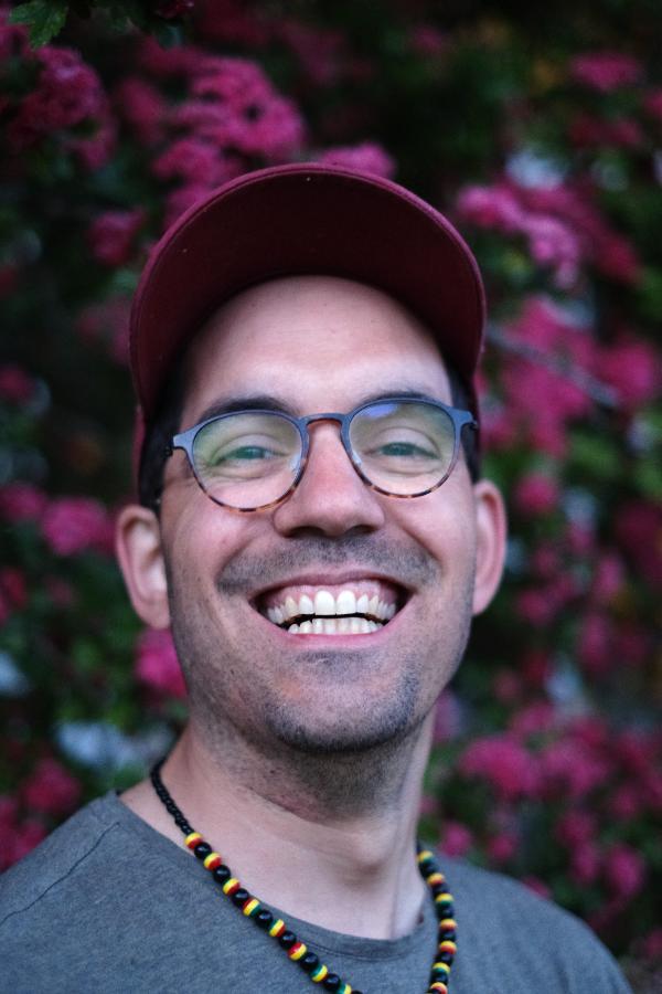 Aarón Blanco Tejedor wearing glasses and a hat smiles in front of a tree full of pink flowers.