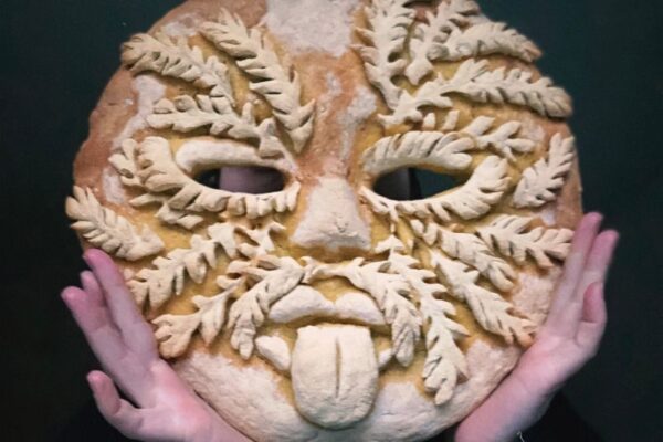 This is a whimsical photo capturing Polina Choni in a unique mask made entirely of bread. The mask is adorned with delicate leaves, adding an intriguing touch to the overall aesthetic.